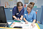 Two healthcare staff members reviewing documents together at a workstation, with one pointing to information on a printed chart.