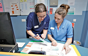 Two healthcare staff members reviewing documents together at a workstation, with one pointing to information on a printed chart.