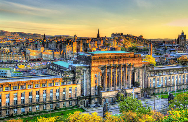 A panoramic view of Edinburgh at golden hour, featuring St Andrew's House building in the foreground and the city skyline with historic architecture and rolling hills in the background.