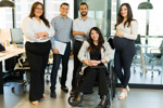 Five people in a modern office, with one person seated in a wheelchair and four others standing nearby. Office desks, computers and meeting rooms are visible in the background.