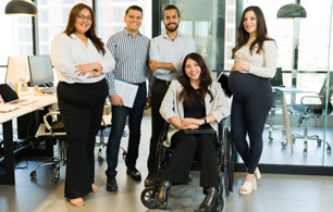Five people in a modern office, with one person seated in a wheelchair and four others standing nearby. Office desks, computers and meeting rooms are visible in the background.