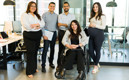 Five people in a modern office, with one person seated in a wheelchair and four others standing nearby. Office desks, computers and meeting rooms are visible in the background.