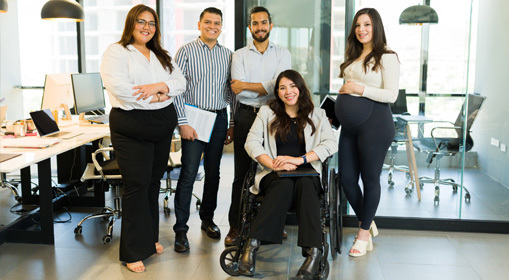 Five people in a modern office, with one person seated in a wheelchair and four others standing nearby. Office desks, computers and meeting rooms are visible in the background.