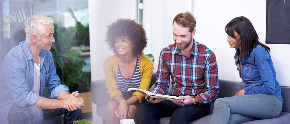 Group of work colleagues in smart casual dress sitting on a sofa against bright background smiling in discussion about an open journal article.