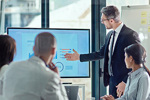 Office setting, four colleagues gathered round a data presentation screen with one colleague pointing at screen and others looking on, flipboard in background