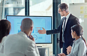 Office setting, four colleagues gathered round a data presentation screen with one colleague pointing at screen and others looking on, flipboard in background