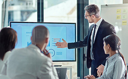 Office setting, four colleagues gathered round a data presentation screen with one colleague pointing at screen and others looking on, flipboard in background