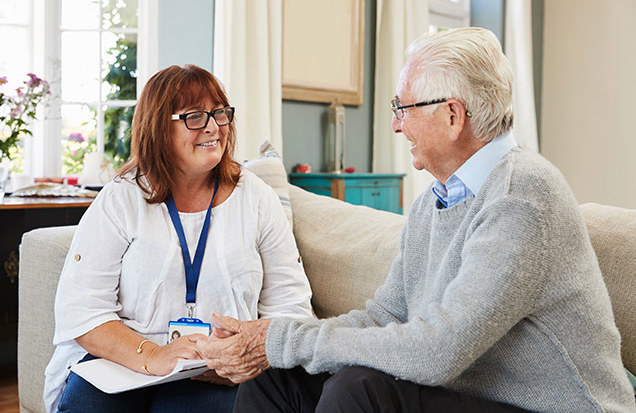 A care professional sits with an older man in a comfortable living room, holding his hand while listening and offering support during a conversation.