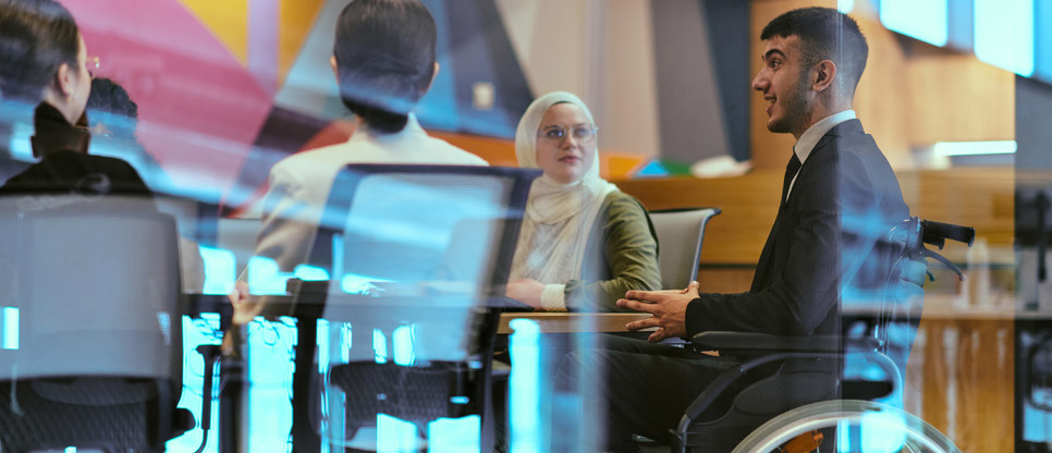 People seated around a meeting table in a modern office, including a wheelchair user, engaged in discussion.
