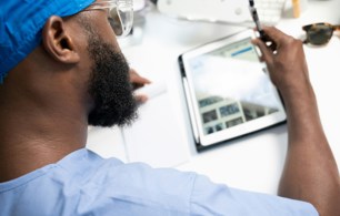 Person in blue clinical scrubs and protective glasses using a tablet computer and stylus at a desk in a healthcare setting.