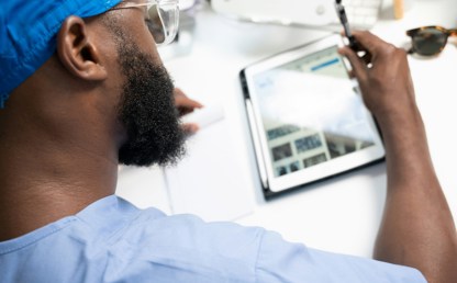 Person in blue clinical scrubs and protective glasses using a tablet computer and stylus at a desk in a healthcare setting.