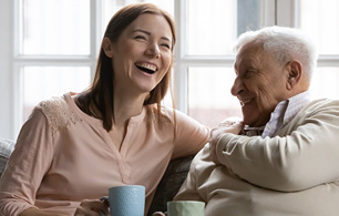 Two people sitting together on a sofa by a window, leaning toward each other and smiling and laughing in a bright indoor setting.
