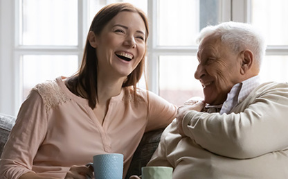 Two people sitting together on a sofa by a window, leaning toward each other and smiling and laughing in a bright indoor setting.