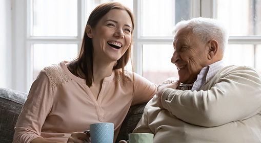 Two people sitting together on a sofa by a window, leaning toward each other and smiling and laughing in a bright indoor setting.