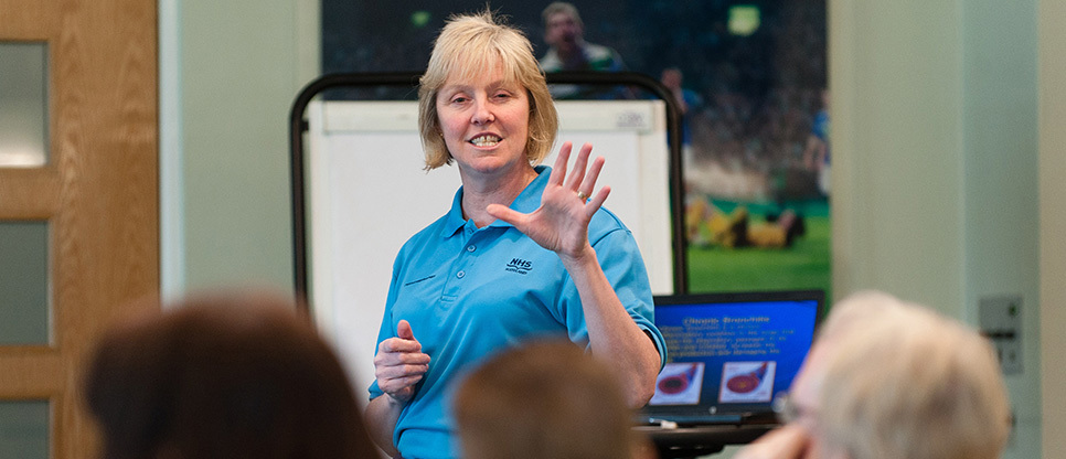Presenter speaking to a seated audience, gesturing with one hand beside a flipchart and presentation screen in a meeting room.