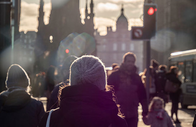 People walk along a busy city street at sunset, with traffic lights, public transport, and historic buildings visible in the background.