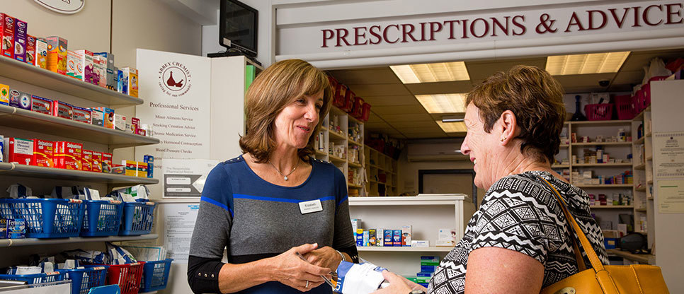 Person handing a prescription item to a customer across the counter in a community pharmacy, with shelves of healthcare products and medicines in the background.