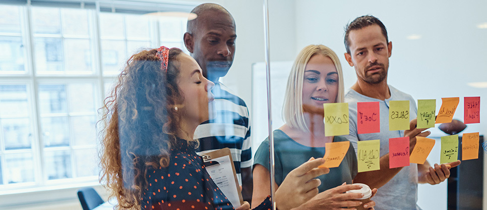 Group of people standing around a glass wall covered with sticky notes, discussing ideas and reviewing notes during a collaborative workshop.