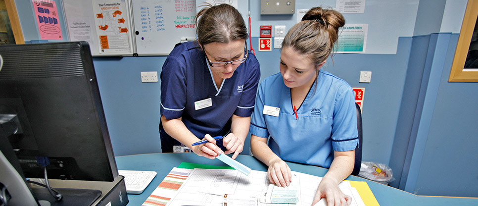 Two healthcare staff members reviewing documents together at a workstation, with one pointing to information on a printed chart.