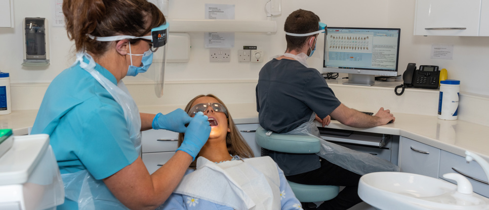 Dentist in protective wear examining a patients teeth, dental nurse seated at desk in background updating digital dental health record on computer screen