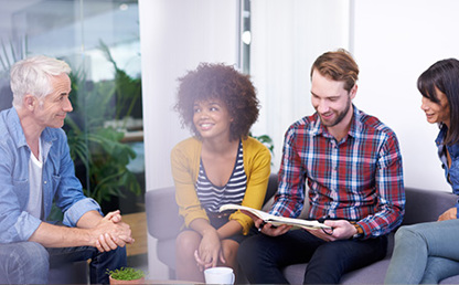 Group of work colleagues in smart casual dress sitting on a sofa against bright background smiling in discussion about an open journal article.
