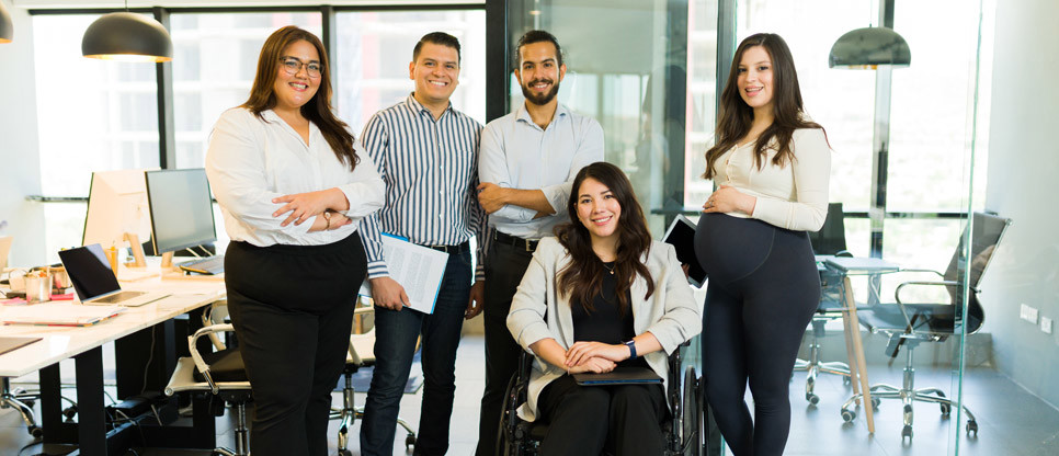 Five people in a modern office, with one person seated in a wheelchair and four others standing nearby. Office desks, computers and meeting rooms are visible in the background.
