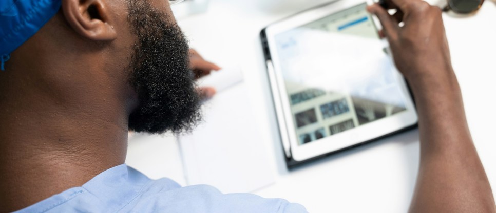 Person in blue clinical scrubs and protective glasses using a tablet computer and stylus at a desk in a healthcare setting.