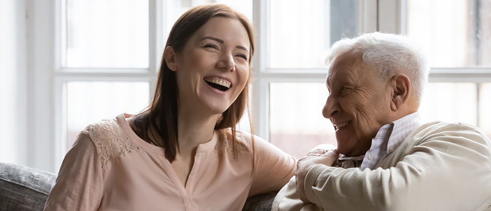 Two people sitting together on a sofa by a window, leaning toward each other and smiling and laughing in a bright indoor setting.