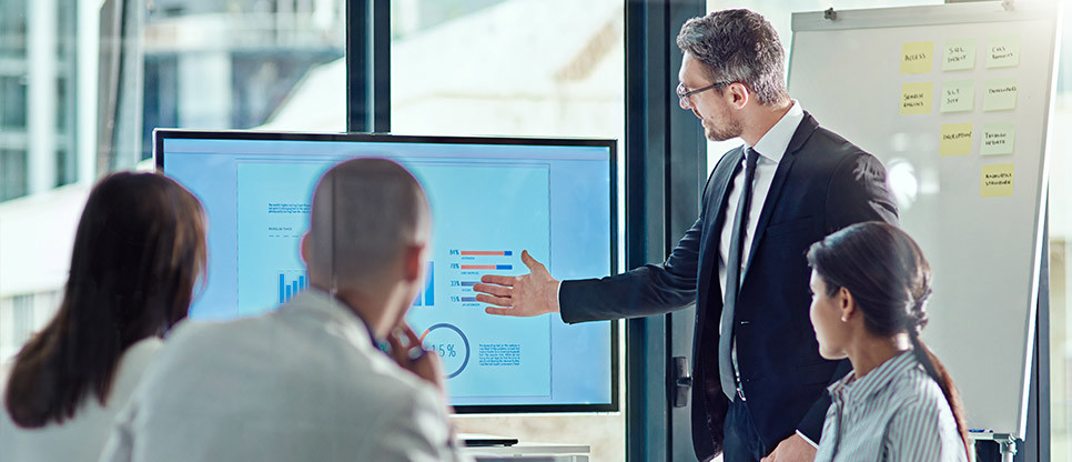Office setting, four colleagues gathered round a data presentation screen with one colleague pointing at screen and others looking on, flipboard in background