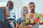 Group of people standing around a glass wall covered with sticky notes, discussing ideas and reviewing notes during a collaborative workshop.