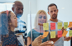 Group of people standing around a glass wall covered with sticky notes, discussing ideas and reviewing notes during a collaborative workshop.
