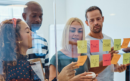 Group of people standing around a glass wall covered with sticky notes, discussing ideas and reviewing notes during a collaborative workshop.