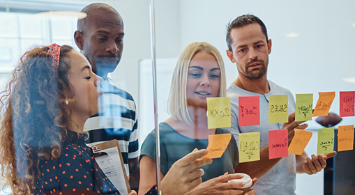 Group of people standing around a glass wall covered with sticky notes, discussing ideas and reviewing notes during a collaborative workshop.