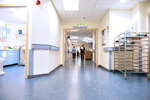A well-lit hospital corridor showing the entrance to a ward, two people walking in the corridor and a person sitting at a desk.