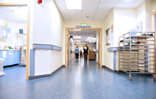 A well-lit hospital corridor showing the entrance to a ward, two people walking in the corridor and a person sitting at a desk.