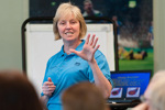 Presenter speaking to a seated audience, gesturing with one hand beside a flipchart and presentation screen in a meeting room.