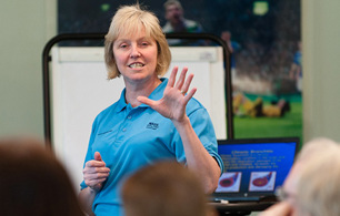 Presenter speaking to a seated audience, gesturing with one hand beside a flipchart and presentation screen in a meeting room.