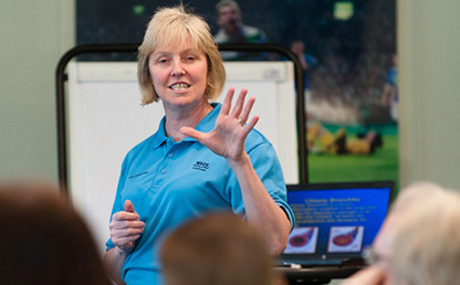 Presenter speaking to a seated audience, gesturing with one hand beside a flipchart and presentation screen in a meeting room.