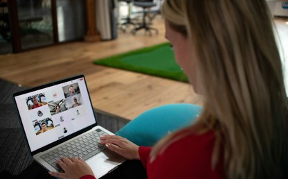 Person sitting on a sofa using a laptop to take part in an online webinar with several participants visible on screen.