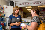 Person handing a prescription item to a customer across the counter in a community pharmacy, with shelves of healthcare products and medicines in the background.