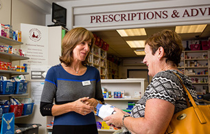 Person handing a prescription item to a customer across the counter in a community pharmacy, with shelves of healthcare products and medicines in the background.