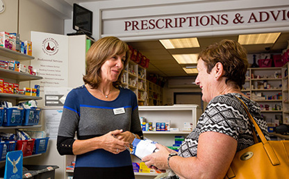 Person handing a prescription item to a customer across the counter in a community pharmacy, with shelves of healthcare products and medicines in the background.