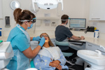 Dentist in protective wear examining a patients teeth, dental nurse seated at desk in background updating digital dental health record on computer screen