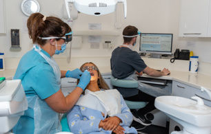 Dentist in protective wear examining a patients teeth, dental nurse seated at desk in background updating digital dental health record on computer screen