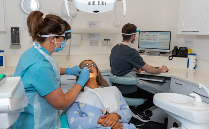 Dentist in protective wear examining a patients teeth, dental nurse seated at desk in background updating digital dental health record on computer screen